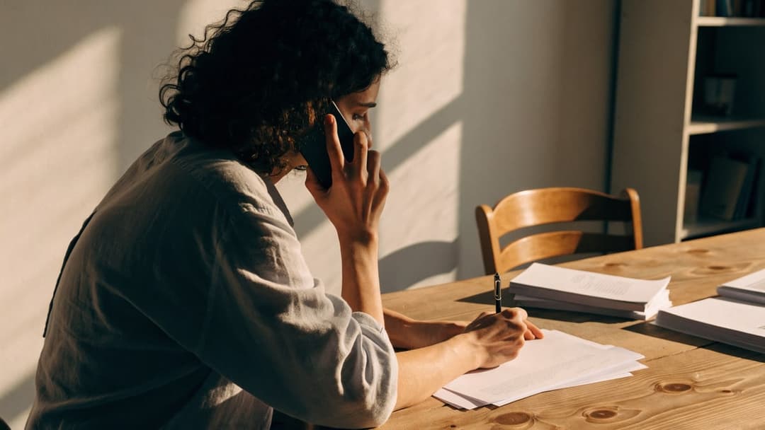 Pessoa ao telefone fazendo anotações em folhas sobre uma mesa.